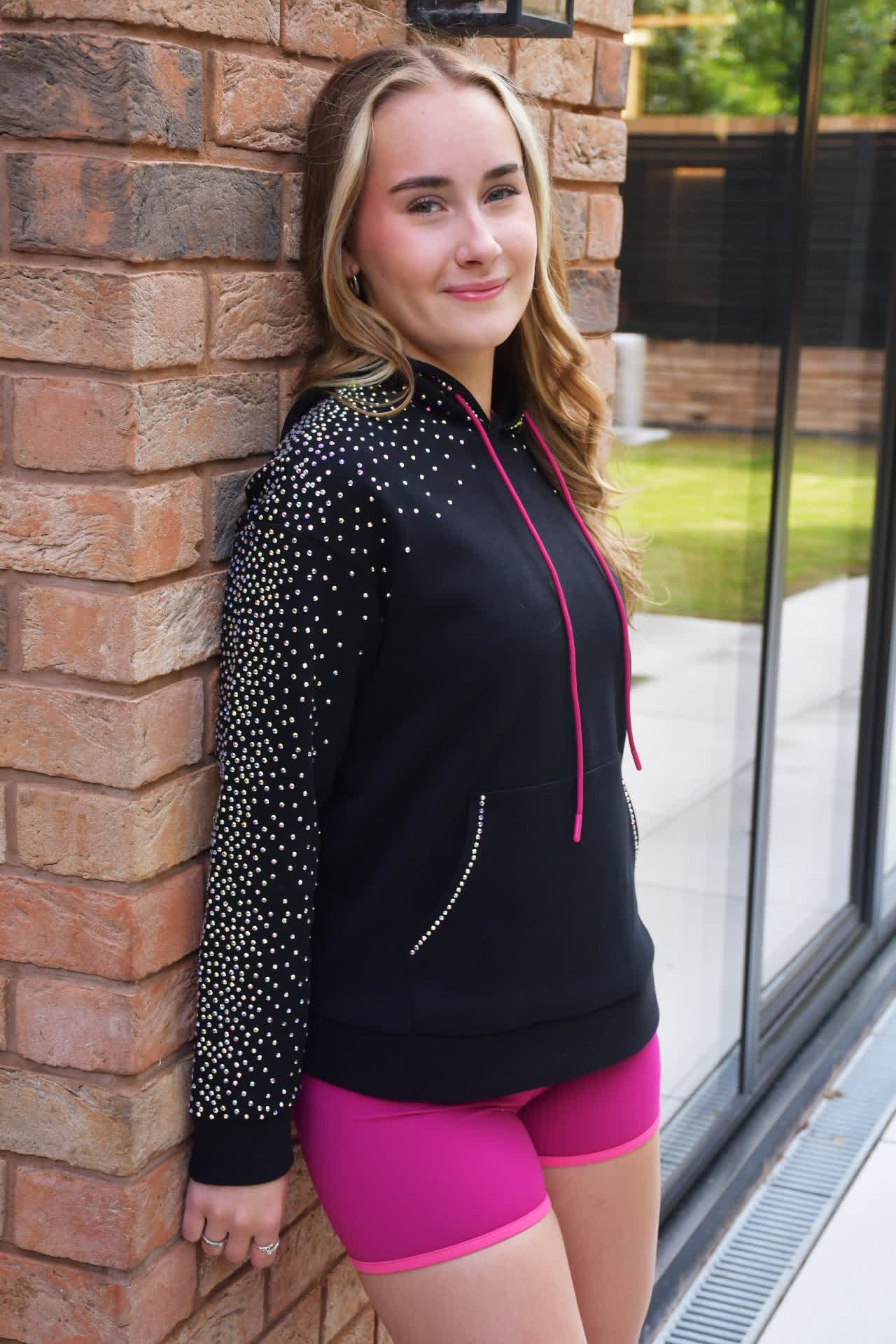 Athlete leaning against a brick wall wearing the black rhinestone Flare Signature Hoodie with pink drawstrings and Mulberry shorts